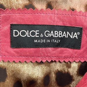 Op de afbeelding: Close-up van de binnenkant van een roze leren kledingstuk, met een zwart label met "DOLCE & GABBANA" en "MADE IN ITALY" in witte tekst. De voering heeft een luipaardprint. Een zilveren ketting is zichtbaar aan de bovenkant.