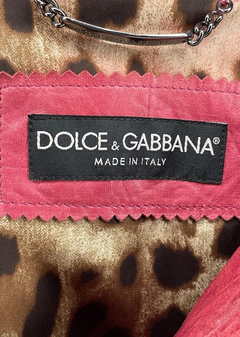 Op de afbeelding: Close-up van de binnenkant van een roze leren kledingstuk, met een zwart label met "DOLCE & GABBANA" en "MADE IN ITALY" in witte tekst. De voering heeft een luipaardprint. Een zilveren ketting is zichtbaar aan de bovenkant.