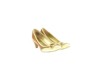 Op de afbeelding: Een paar tweekleurige pumps met een lage hak. De schoenen zijn bruin en crèmekleurig met een strikdetail op de neus. De hakken zijn ongeveer 5 cm hoog. De schoenen hebben een klassiek, elegant ontwerp.