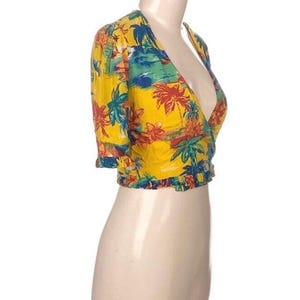 Puede incluir: Un top envolvente amarillo con un estampado tropical con palmeras, flores y una escena de playa. El top de manga corta tiene un dobladillo con volantes y se muestra en un maniqu&iacute;.