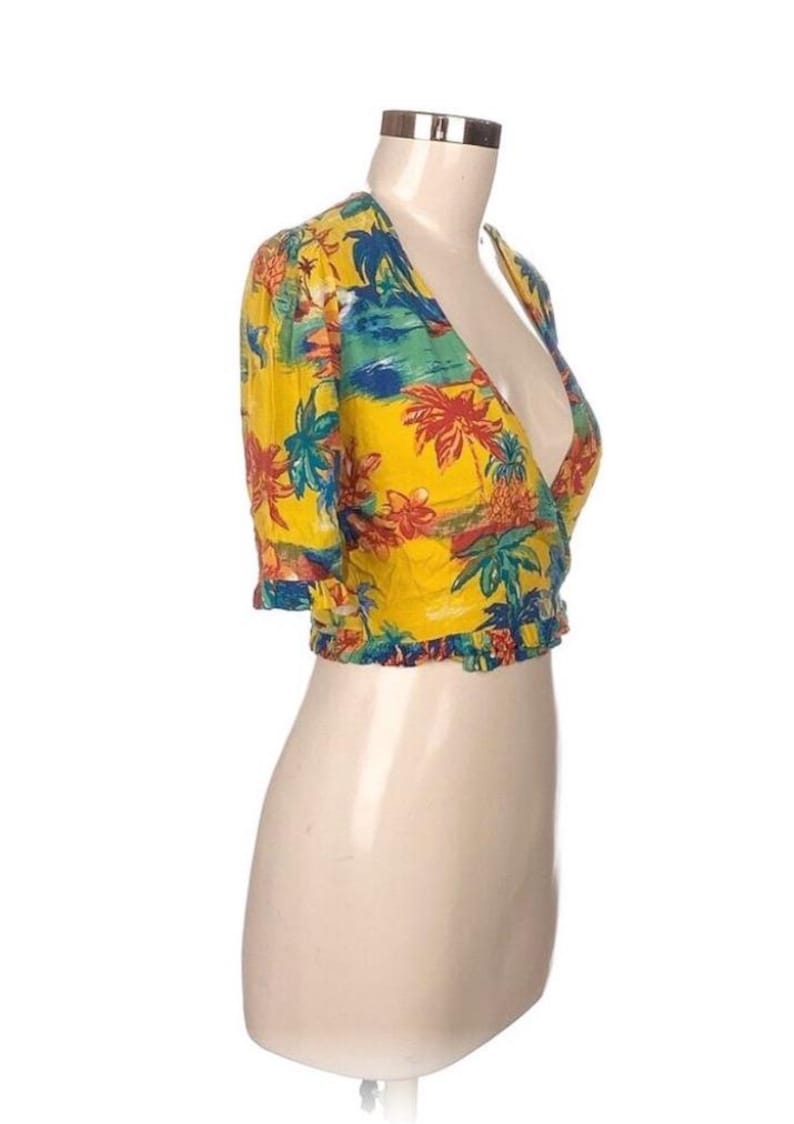 Puede incluir: Un top envolvente amarillo con un estampado tropical con palmeras, flores y una escena de playa. El top de manga corta tiene un dobladillo con volantes y se muestra en un maniqu&iacute;.