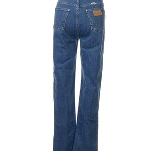 May include: Blue denim jeans with a classic five-pocket design. The jeans have a straight leg cut and a leather patch on the back pocket. The Wrangler logo is visible on the patch. The jeans are displayed against a white background.