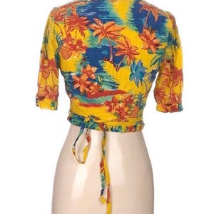 Puede incluir: Un top envolvente amarillo con un estampado tropical con palmeras, flores y pi&ntilde;as en tonos rojos, azules y naranjas. El top tiene mangas cortas y un cierre de lazo en la cintura. La prenda se exhibe en un maniqu&iacute;.