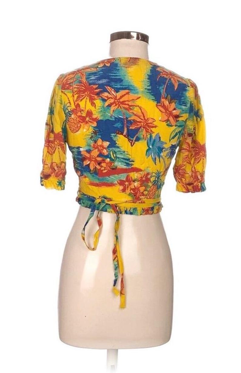 Puede incluir: Un top envolvente amarillo con un estampado tropical con palmeras, flores y pi&ntilde;as en tonos rojos, azules y naranjas. El top tiene mangas cortas y un cierre de lazo en la cintura. La prenda se exhibe en un maniqu&iacute;.