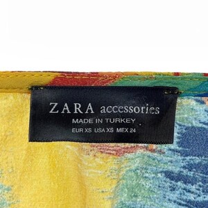 Puede incluir: Primer plano de una etiqueta de ZARA accessories. La etiqueta negra muestra texto blanco: "ZARA accessories MADE IN TURKEY EUR XS USA XS MEX 24". La tela presenta un colorido patr&oacute;n abstracto con tonos amarillos, azules, rojos y verdes.