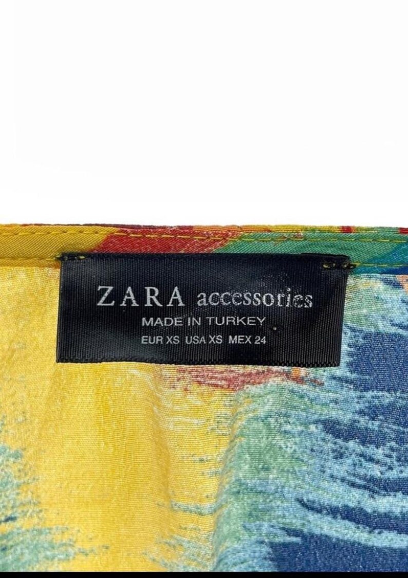 Puede incluir: Primer plano de una etiqueta de ZARA accessories. La etiqueta negra muestra texto blanco: "ZARA accessories MADE IN TURKEY EUR XS USA XS MEX 24". La tela presenta un colorido patr&oacute;n abstracto con tonos amarillos, azules, rojos y verdes.