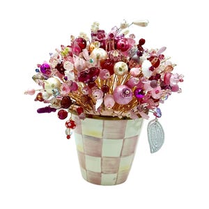 May include: A decorative floral arrangement featuring a variety of pink, red, and white beads and baubles. The arrangement is set in a checkered vase with a silver leaf accent. The beads are arranged on gold wire.
