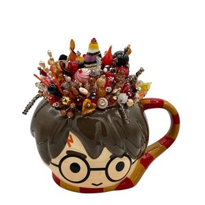Pode incluir: Uma caneca de cerâmica em forma de personagem com cabelo castanho, óculos pretos redondos e um símbolo de raio na testa. A caneca está cheia com um arranjo decorativo de várias contas e ornamentos de vidro em tons de vermelho, laranja e castanho.