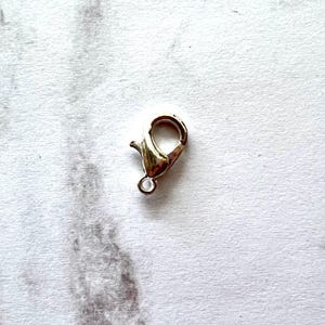 May include: A gold-colored lobster clasp, a jewelry component, is shown against a white background. The clasp has a spring-loaded mechanism for secure closure and a small loop for attaching to other jewelry pieces.