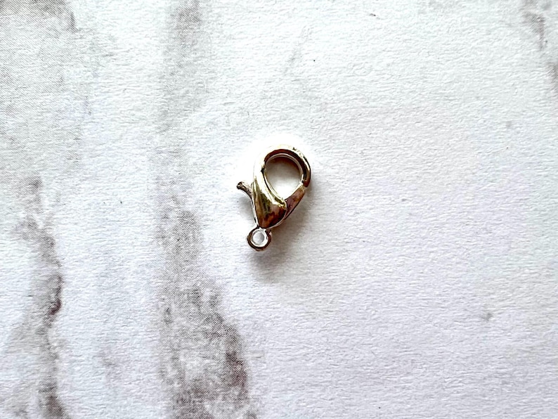 May include: A gold-colored lobster clasp, a jewelry component, is shown against a white background. The clasp has a spring-loaded mechanism for secure closure and a small loop for attaching to other jewelry pieces.