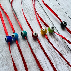 May include: Seven red satin cords with different coloured evil eye beads. The beads are blue, turquoise, green, yellow, black, and white.