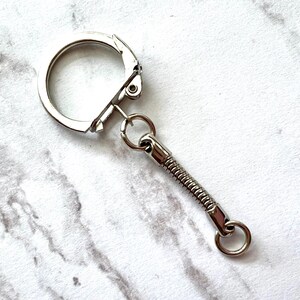 May include: A silver-colored keychain with a spring-loaded extension. The keychain has a large, round clasp and a small ring at the end of the extension. The background is a white and gray marble pattern.