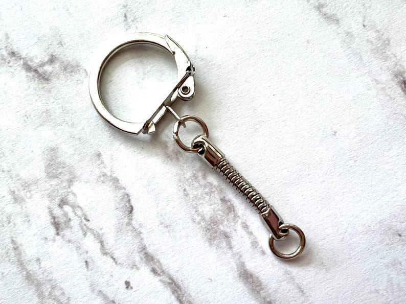 May include: A silver-colored keychain with a spring-loaded extension. The keychain has a large, round clasp and a small ring at the end of the extension. The background is a white and gray marble pattern.
