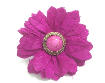 May include: A large, pink fabric flower with a gold-toned button in the center. The button has a pink stone with white marbling.