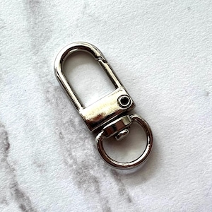 May include: A shiny, silver-colored swivel clasp with a D-ring. The clasp has a rectangular body with a curved top and a small, circular ring attached at the bottom. The metal hardware is designed for attaching to bags or other accessories.
