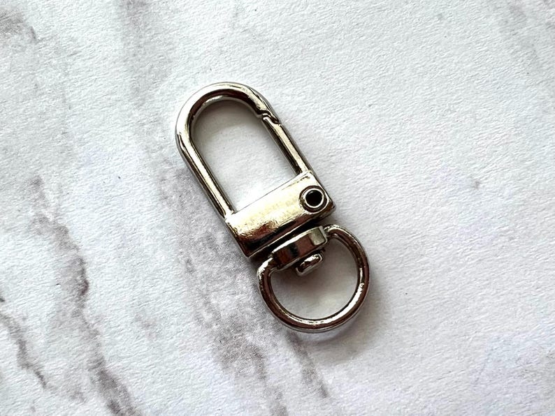 May include: A shiny, silver-colored swivel clasp with a D-ring. The clasp has a rectangular body with a curved top and a small, circular ring attached at the bottom. The metal hardware is designed for attaching to bags or other accessories.