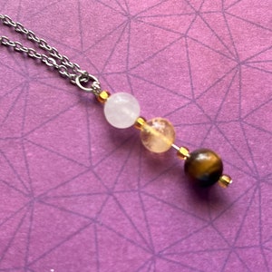 May include: A silver chain necklace with three gemstone beads: a white, a yellow, and a brown bead. The beads are strung together with small gold-colored beads. The necklace is displayed on a purple surface.