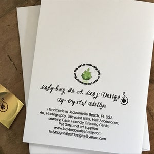 May include: A white card with a green leaf design and the text "This card is made using 30% of post consumer waste."  The text "Lady bug, On A Leaf Design" is written in black cursive script. Below the cursive text is the text "By Crystal Phillips" in black script.  The text "Handmade in Jacksonville Beach, FL USA" is written in black.  Below the text is a list of items: "Art, Photography, Upcycled Gifts, Hair Accessories, Jewelry, Earth Friendly Greeting Cards, Pet Gifts and art supplies."  The text "www.ladybugonaleaf.etsy.com" and "ladybugonaleafdesigns@yahoo.com" are written in black.