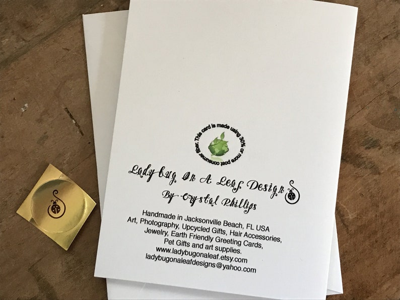 May include: A white card with a green leaf design and the text "This card is made using 30% of post consumer waste."  The text "Lady bug, On A Leaf Design" is written in black cursive script. Below the cursive text is the text "By Crystal Phillips" in black script.  The text "Handmade in Jacksonville Beach, FL USA" is written in black.  Below the text is a list of items: "Art, Photography, Upcycled Gifts, Hair Accessories, Jewelry, Earth Friendly Greeting Cards, Pet Gifts and art supplies."  The text "www.ladybugonaleaf.etsy.com" and "ladybugonaleafdesigns@yahoo.com" are written in black.