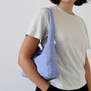 Può includere: Una borsa a tracolla all'uncinetto, blu chiaro, di forma rettangolare e con una lunga tracolla. La borsa è realizzata con un filato strutturato. La borsa è tenuta da una persona che indossa una maglietta bianca e pantaloni neri.