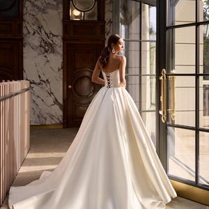 May include: A white satin wedding dress with a corset back and a full skirt. The dress is displayed in a room with a marble wall and a large window. The number 220 is visible in the top right corner of the image.