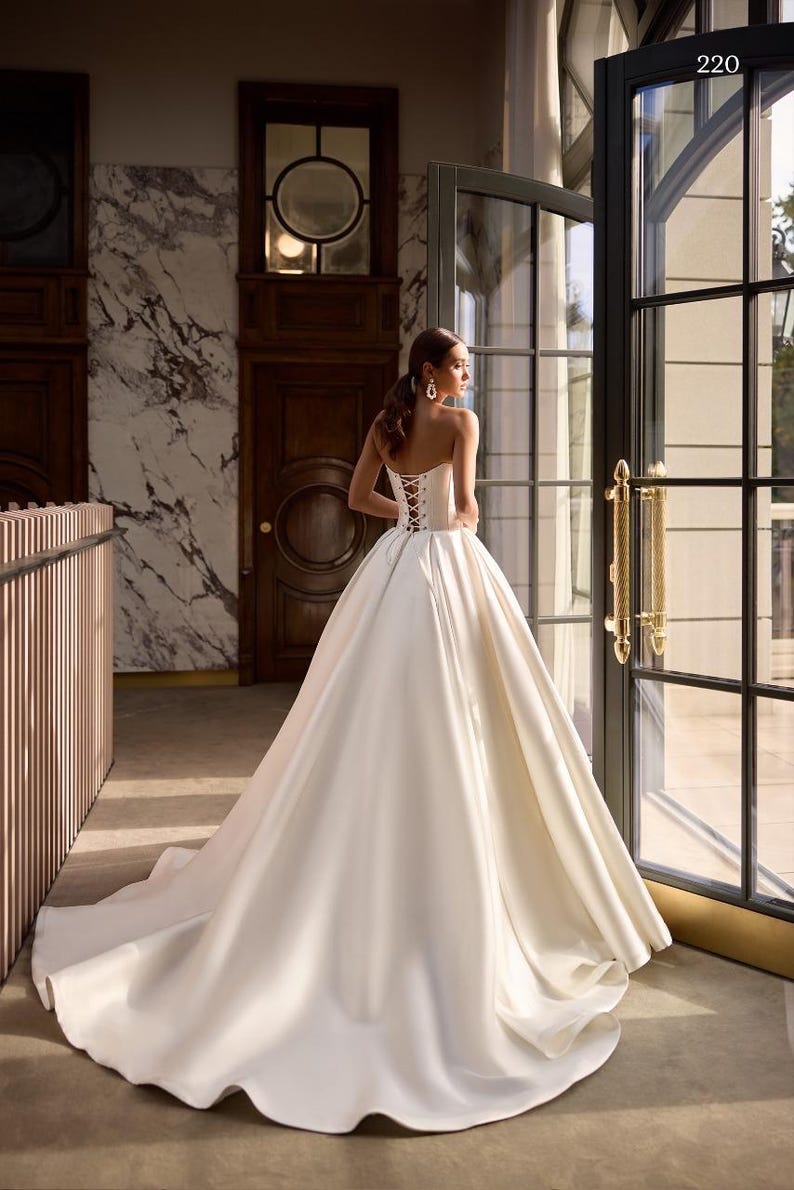 May include: A white satin wedding dress with a corset back and a full skirt. The dress is displayed in a room with a marble wall and a large window. The number 220 is visible in the top right corner of the image.