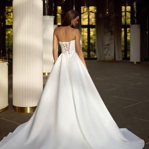 May include: A white strapless wedding dress with a lace-up corset back. The dress has a full skirt and a train. The dress is displayed on a model in a large, empty room with white columns.