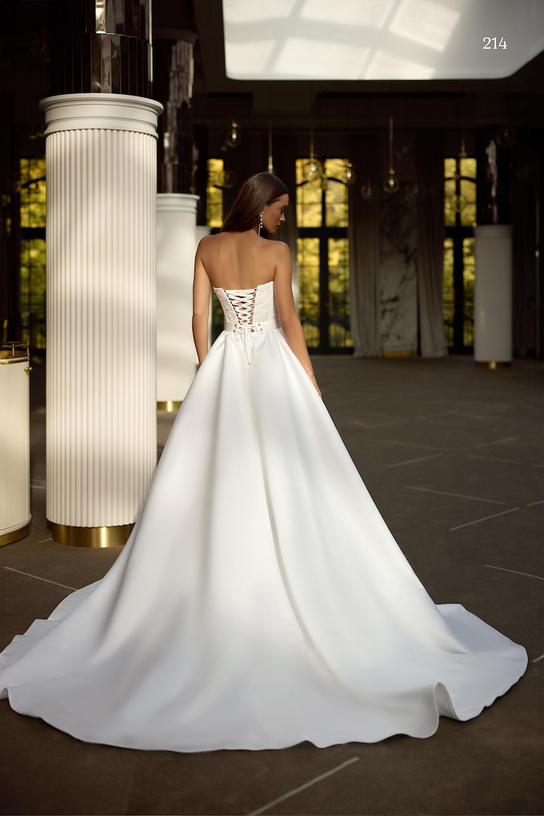 May include: A white strapless wedding dress with a lace-up corset back. The dress has a full skirt and a train. The dress is displayed on a model in a large, empty room with white columns.