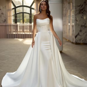 May include: A white strapless wedding dress with a fitted bodice and a flowing skirt. The dress has a delicate lace overlay on the bodice and a satin skirt. The dress is shown on a model in a luxurious setting.