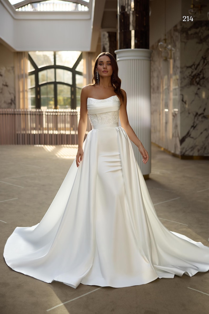 May include: A white strapless wedding dress with a fitted bodice and a flowing skirt. The dress has a delicate lace overlay on the bodice and a satin skirt. The dress is shown on a model in a luxurious setting.
