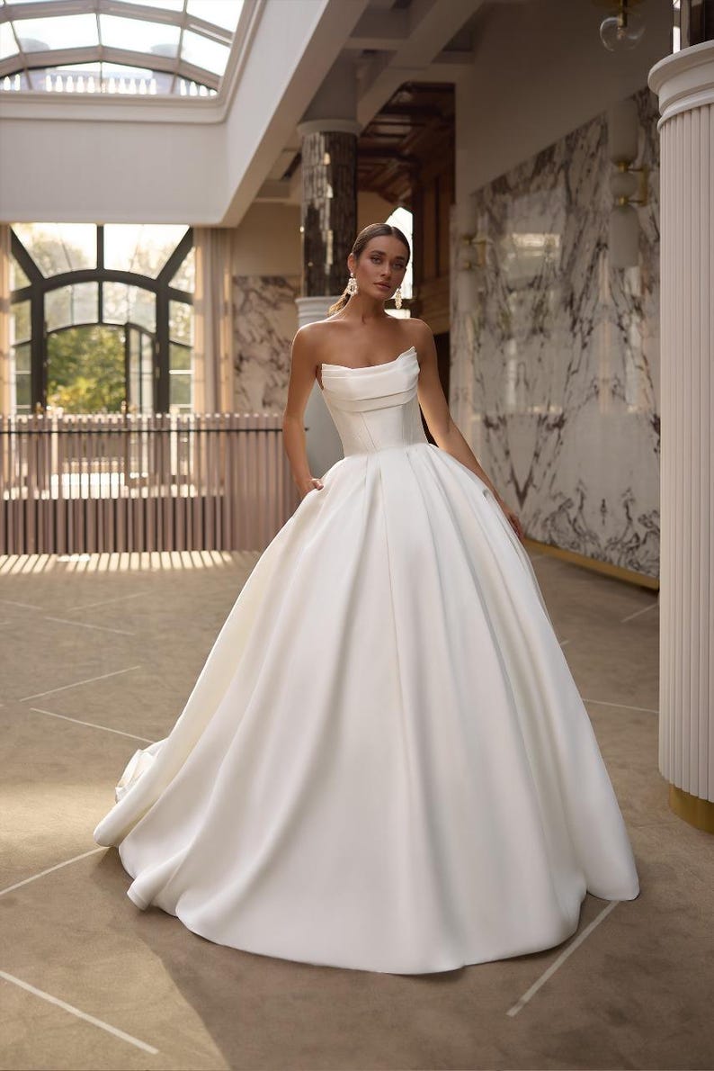 May include: A white strapless wedding dress with a fitted bodice and a full, flowing skirt. The dress is made of a smooth, satin fabric and has a simple, elegant design.