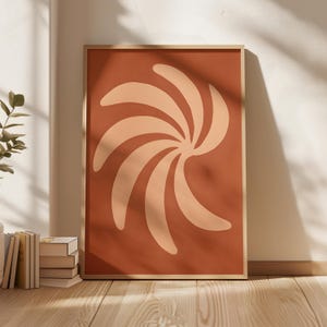 Op de afbeelding: Een ingelijste abstracte kunstprint met een wervelend ontwerp in perzik- en crèmetinten tegen een terracotta achtergrond. Het kunstwerk wordt gepresenteerd in een licht houten frame, leunend tegen een witte muur.