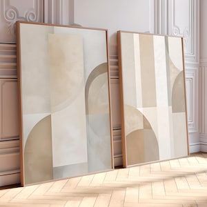 Op de afbeelding: Twee ingelijste abstracte kunstprints. Het kunstwerk toont geometrische vormen in neutrale tinten beige, crème en bruin. De lijsten zijn warmbruin en de prints worden gepresenteerd tegen een witte muur en een houten vloer.