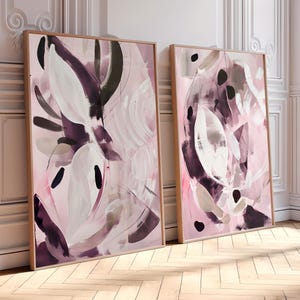 Op de afbeelding: Twee ingelijste abstracte kunstprints. Het kunstwerk toont een mix van roze, witte en donkerpaarse penseelstreken. De lijsten zijn lichtbruin van kleur en de kunst leunt tegen een witte muur en een houten vloer.