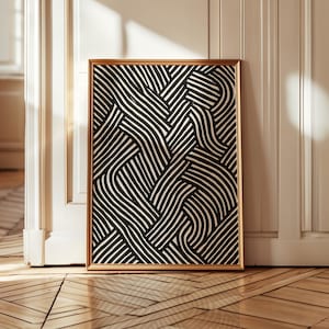 Op de afbeelding: Een ingelijst kunstwerk met een goudkleurige lijst, met een zwart-wit abstract ontwerp. Het kunstwerk wordt getoond tegen een witte muur en een houten vloer. Het abstracte patroon bestaat uit gebogen, verweven lijnen.