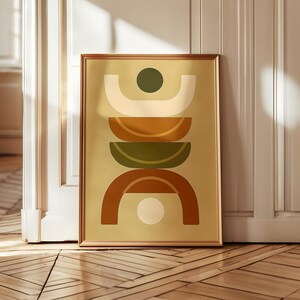 Op de afbeelding: Een ingelijste abstracte kunstprint met een geometrisch ontwerp. Het kunstwerk toont gebogen vormen en cirkels in crème, olijfgroen en roestoranje tegen een lichtgele achtergrond. De lijst heeft een warme bruine kleur.