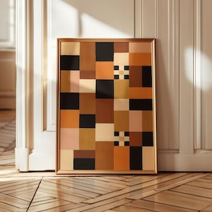 May include: A framed abstract art print featuring a geometric pattern of squares in shades of brown, black, beige, and orange. The artwork has a gold-colored frame and is leaning against a white door.