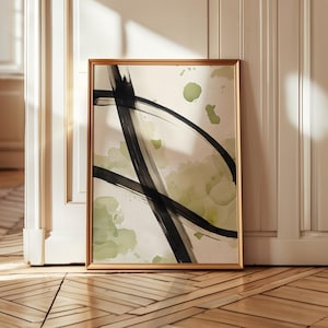 Op de afbeelding: Abstracte kunstafdruk in een gouden lijst. Het kunstwerk toont gedurfde zwarte penseelstreken en zachte, aquarelachtige groene spetters op een crèmekleurige achtergrond. De ingelijste kunst wordt tentoongesteld tegen een witte deur en een houten vloer.