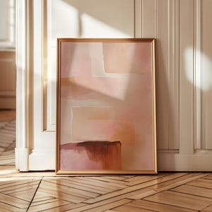 Op de afbeelding: Abstracte kunstafdruk in een gouden lijst. Het kunstwerk toont zachte roze, perzik en bruine penseelstreken. De ingelijste kunst leunt tegen een witte deur en een houten vloer.