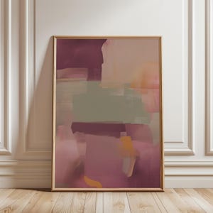 May include: Abstract art print in a wooden frame. The artwork features overlapping brushstrokes in shades of burgundy, pink, peach, and olive green. The framed art is displayed against a white wall and wooden floor.