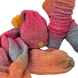 May include: A knitted hat, scarf, and leg warmers set in a gradient of pink, orange, and blue. The hat has a yellow pom-pom and a small tag. The scarf and leg warmers are also knitted and feature the same colour gradient.