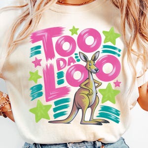 May include: Cream-colored t-shirt featuring a cartoon kangaroo with the text "Too Da Loo" in pink and teal. The design includes green and pink stars. The kangaroo is tan and yellow.