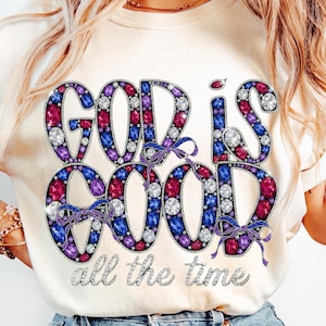 Puede incluir: Camiseta color crema con las palabras "GOD IS GOOD" en un diseño de joyas, con gemas rosas, moradas, azules y transparentes. La frase "all the time" está escrita debajo en una fuente cursiva.