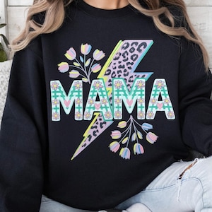 Peut inclure: Sweat-shirt noir avec le mot "MAMA" dans un motif floral et à carreaux. Un éclair jaune, rose et imprimé léopard est derrière le mot. Des accents floraux roses, jaunes et bleus entourent le mot.