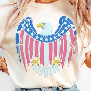 May include: A cream-coloured t-shirt with a patriotic eagle design. The eagle's wings are decorated with blue stars and red and pink stripes, with a light blue head and yellow beak and talons. The design is centered on the shirt.