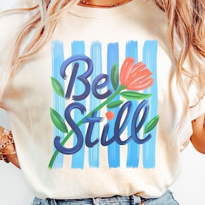 Pode incluir: Camiseta creme com as palavras "Be Still" em escrita azul, acompanhada por uma flor laranja estilizada e folhas verdes. O design é definido contra um fundo de pinceladas azuis verticais.