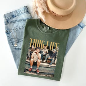May include: An olive green t-shirt with the text "THUG LIFE" and a graphic of four women sitting on a porch. The shirt is paired with light wash denim jeans, a tan hat, and a gold necklace.