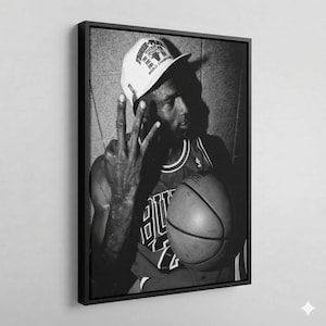 A black and white photograph of a basketball player wearing a Chicago Bulls jersey and a white cap, holding a basketball and making a peace sign gesture.