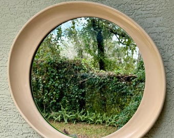 May include: A large, round, cream-colored mirror with a thick frame. The mirror reflects a view of green trees and foliage.