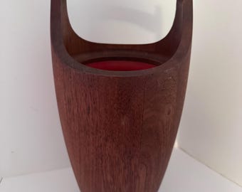 May include: A vintage wooden ice bucket with a handle. The bucket is a deep reddish-brown color, with a smooth, polished finish. The interior is lined with red fabric. The handle is arched and attached to the sides of the bucket.
